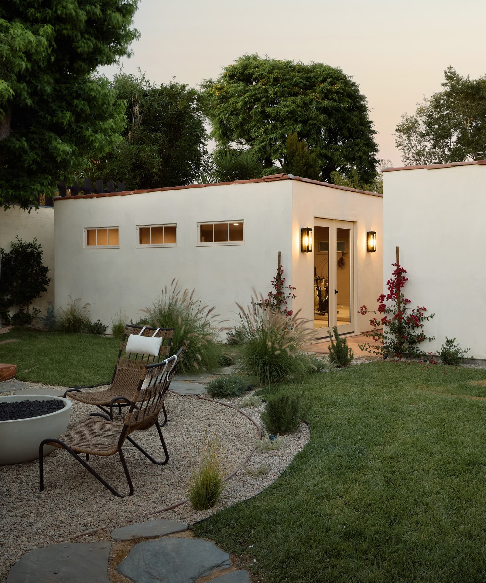 Rear garden at dusk with a fire pit, gravel terrace, lawn and the lit detached ADU