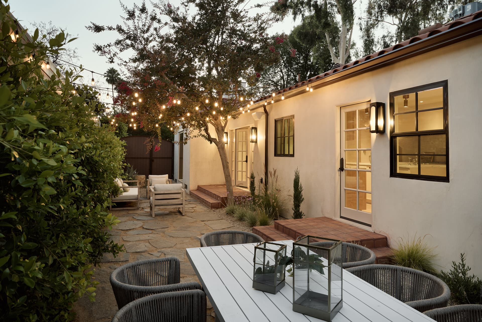 Side courtyard at dusk with string lights, flagstone patio, dining set and warm-lit French doors