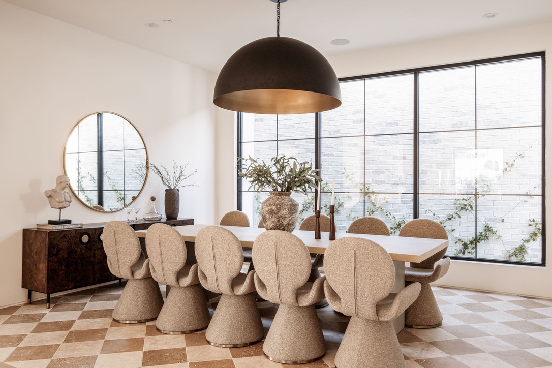Sunlit dining room with a round brass mirror, burl credenza and steel windows onto a white brick courtyard
