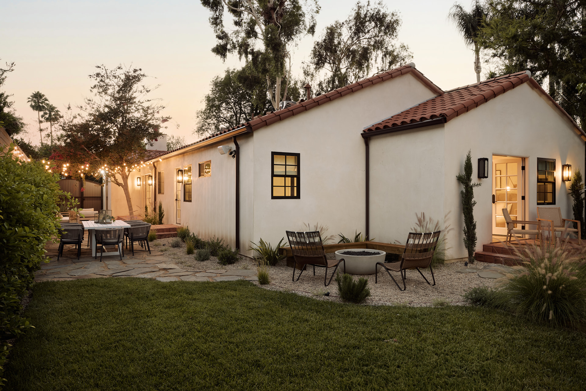 Spanish-style single-storey residence at dusk with clay tile roof, warm lit windows, fire pit lounge and string-lit patio