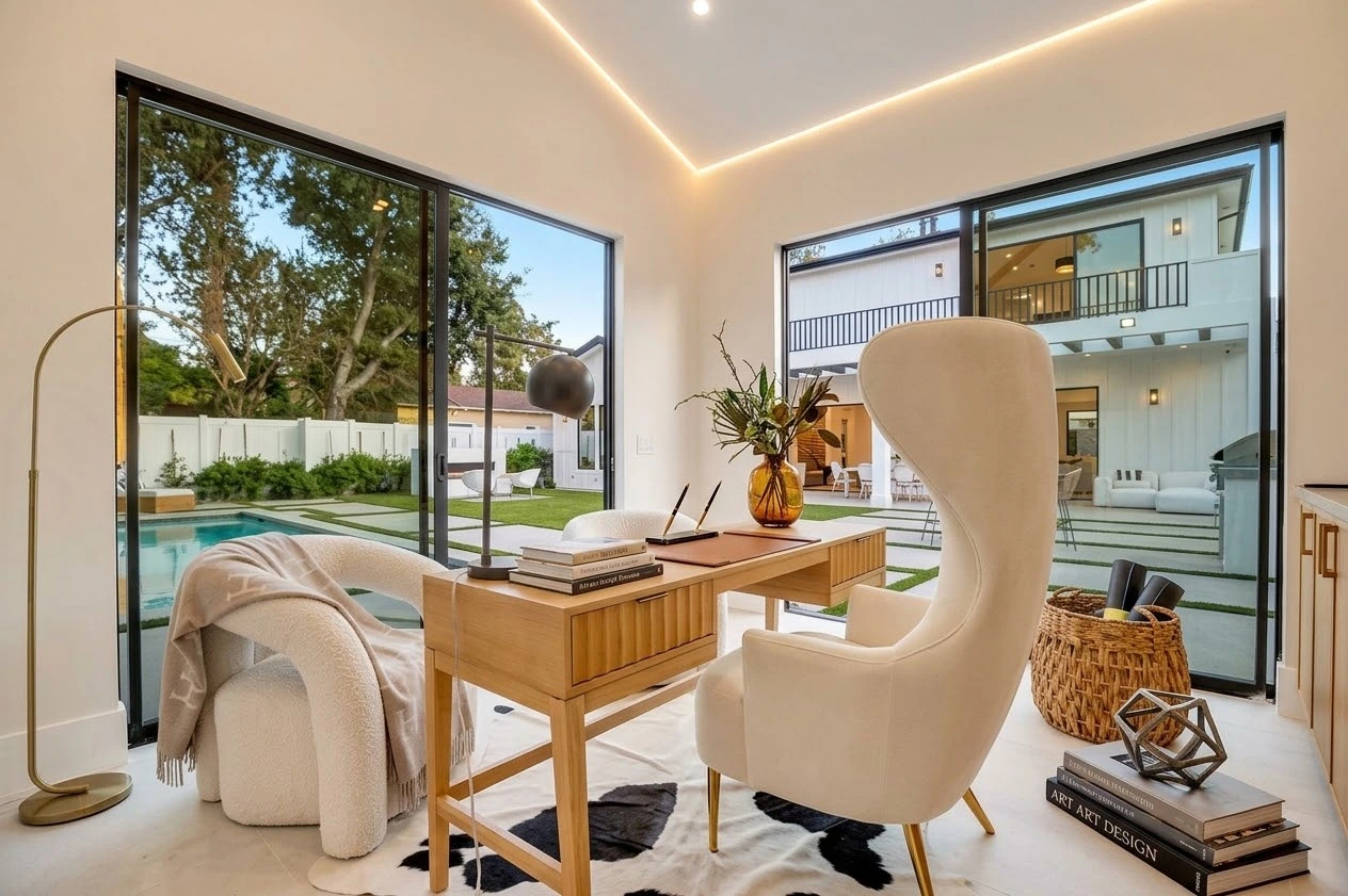 Garden-facing studio with an oak desk, sculptural cream chairs and black-framed windows overlooking the pool