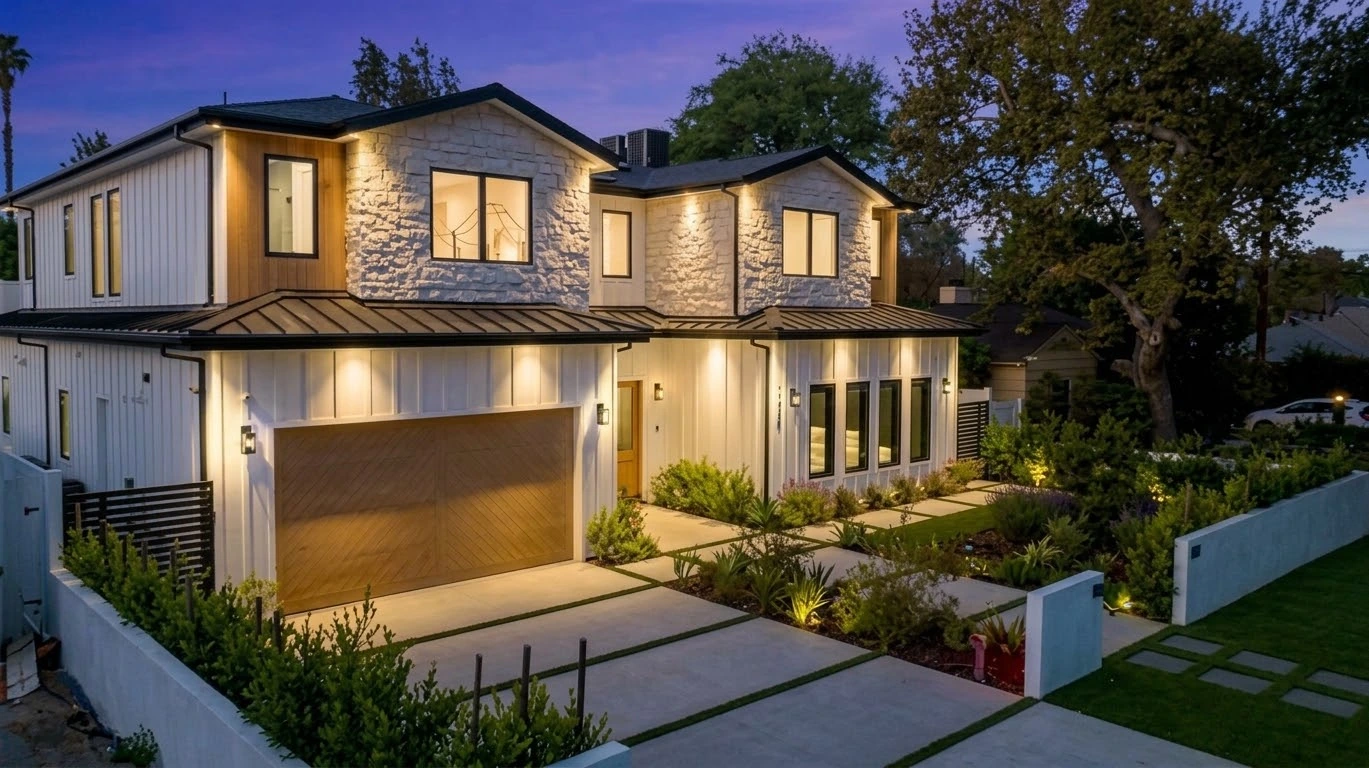 Front elevation at dusk with white siding, stone gables, an oak garage door and landscaped entry