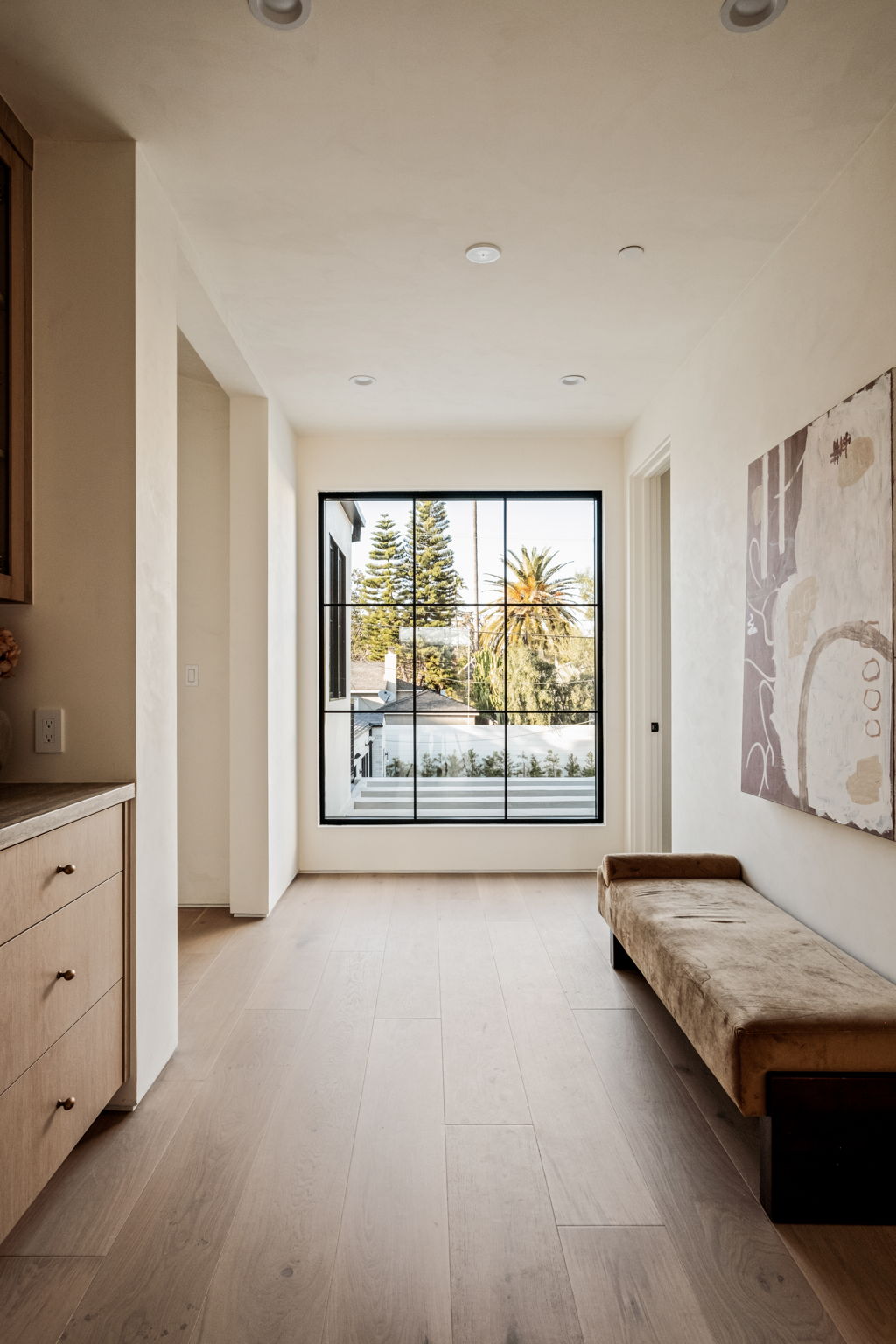 Upper hallway with an oversized steel window, oak cabinetry and a velvet bench beneath abstract art