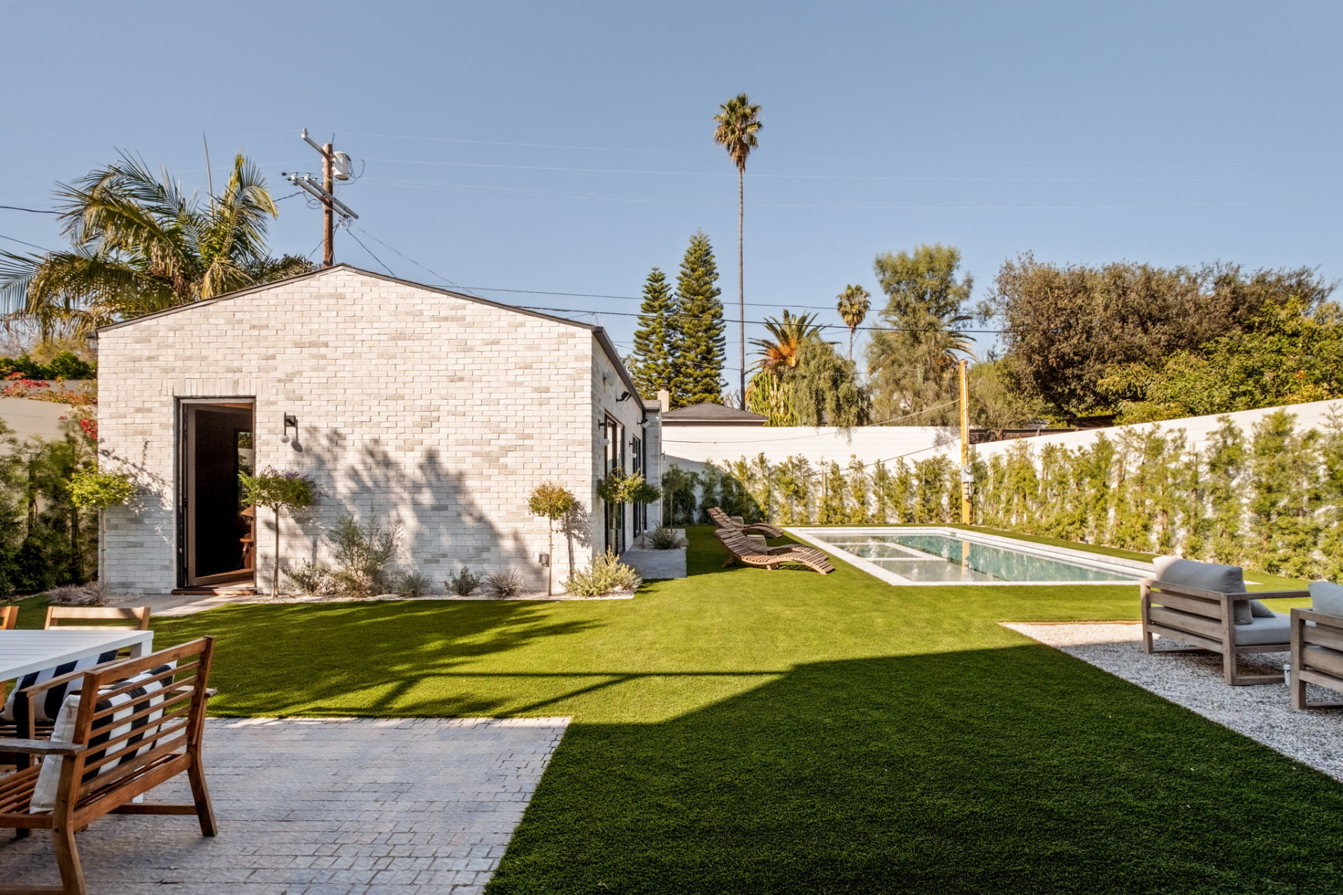 Side view of the white brick ADU with an open steel door, olive topiaries, lawn and the lap pool