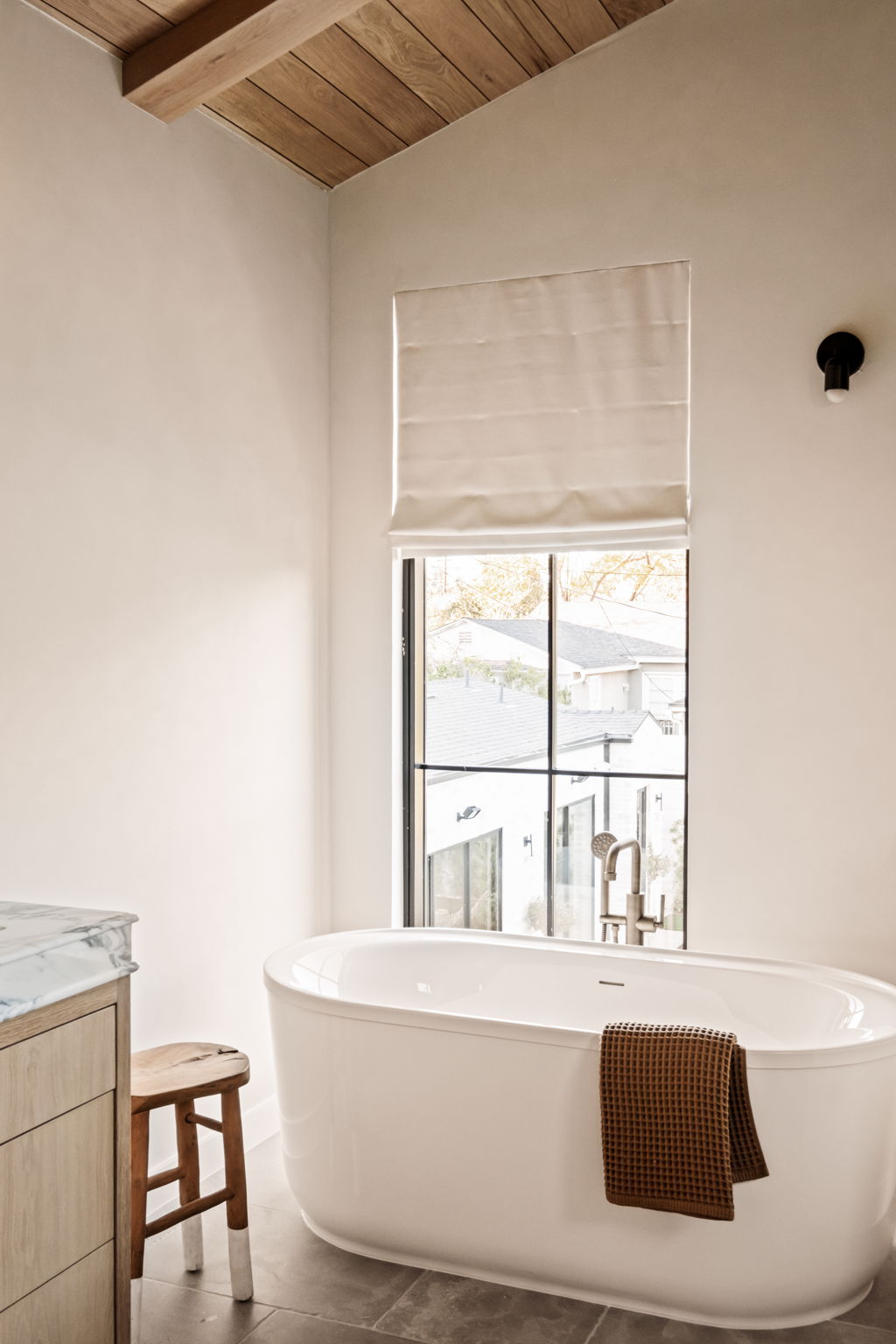 Freestanding soaking tub beneath a linen roman shade and steel window
