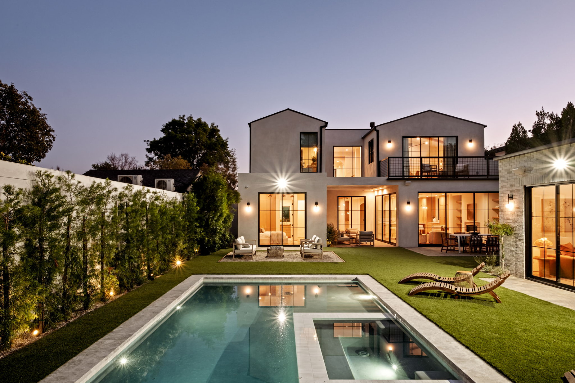 Backyard at twilight with a rectangular pool, spa, lawn and the warm-lit residence beyond