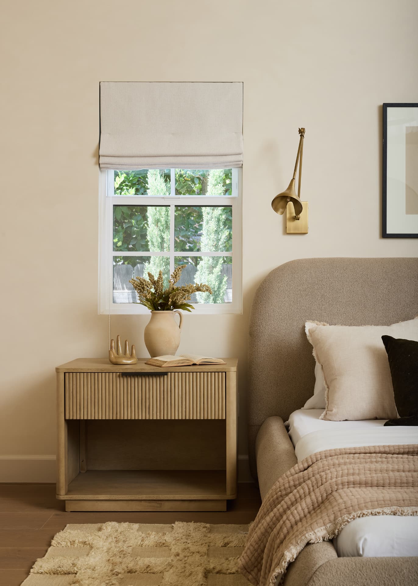 Primary suite with a bouclé bed, fluted oak nightstand and brass sconce beside a linen roman shade