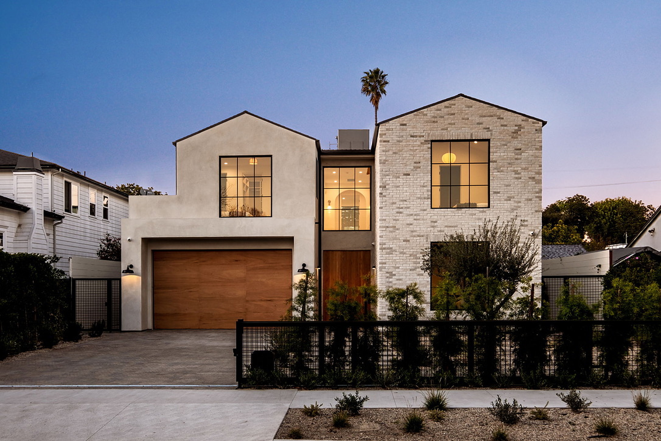 Modern two-storey home at dusk with white brick, smooth stucco gables, black steel windows and an oak garage door
