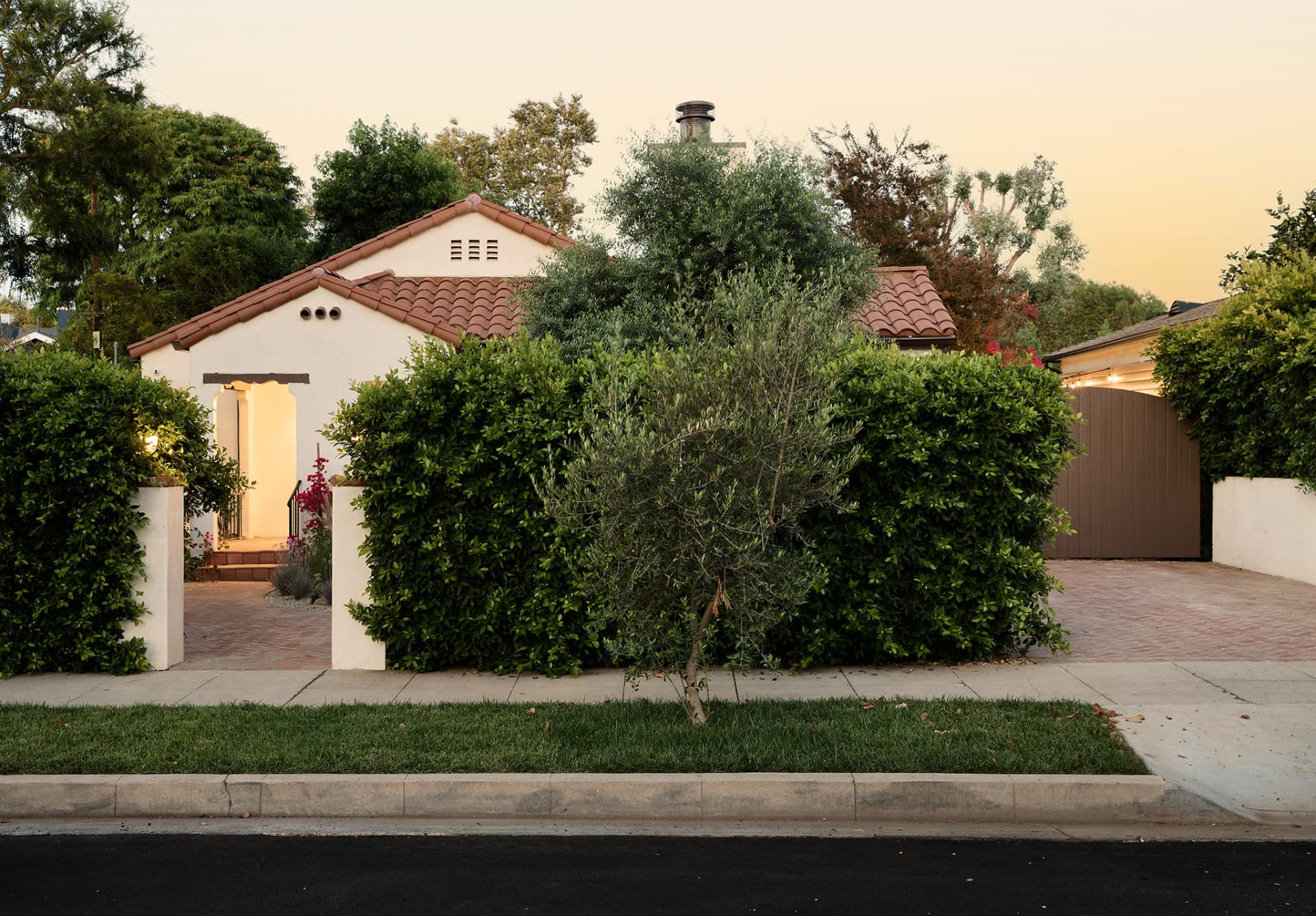 Front facade at sunset with tile roofs, hedges, olive tree and a brick driveway