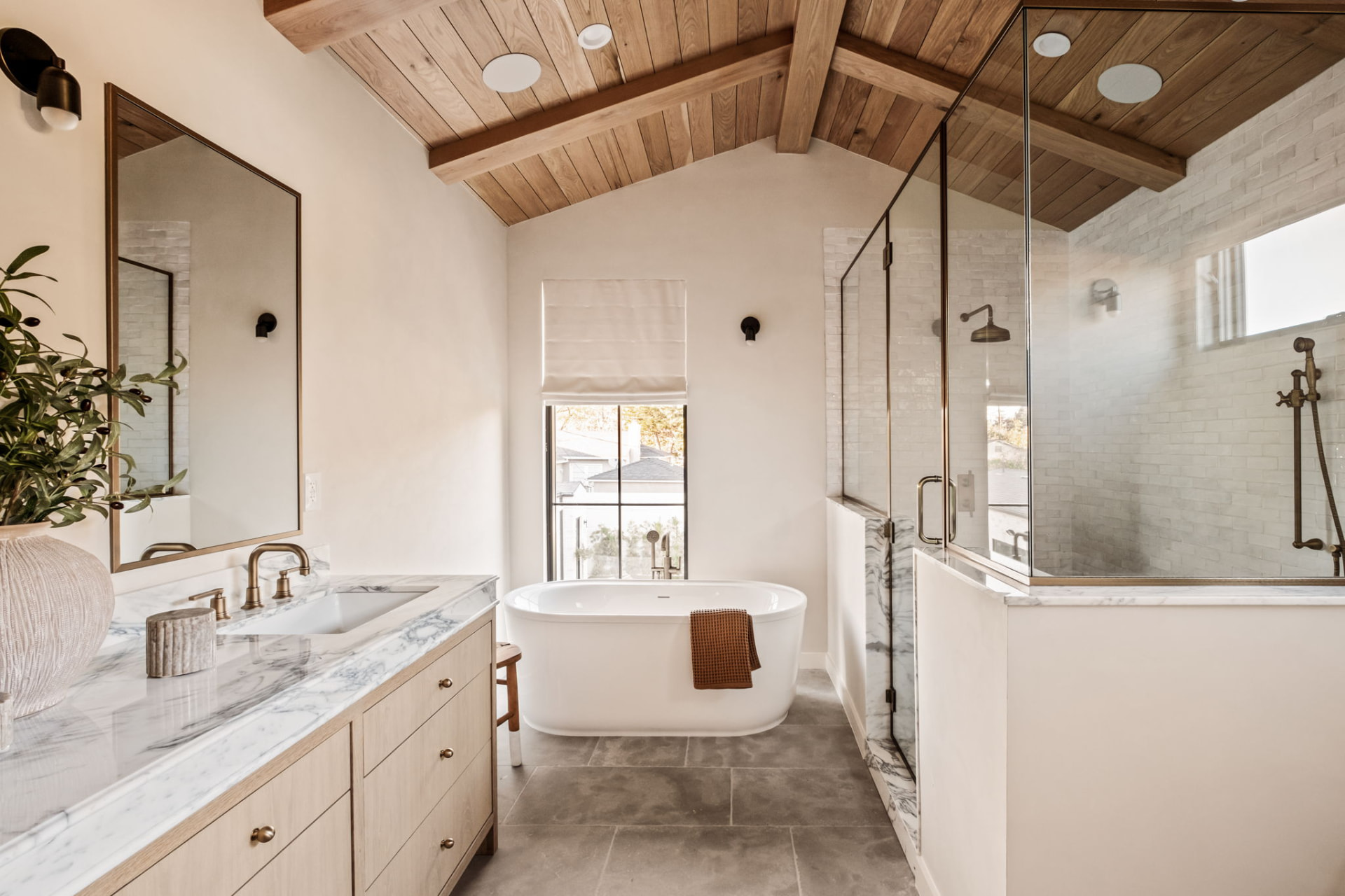 Primary bathroom with a vaulted white oak plank ceiling, marble double vanity, freestanding tub and glass-enclosed brick-tiled shower