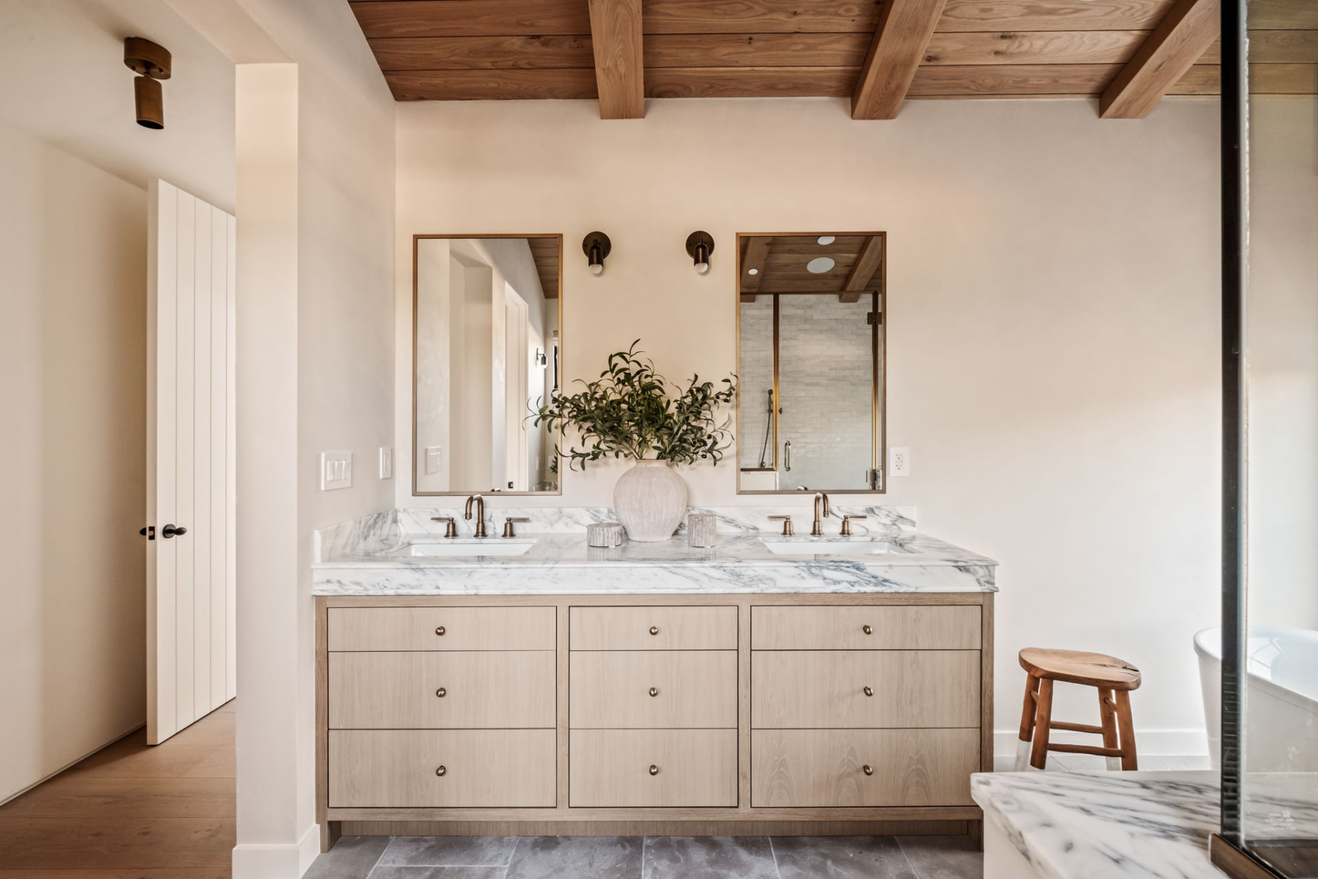 Double marble vanity with brass fixtures and slim mirrors beneath an exposed oak beam ceiling