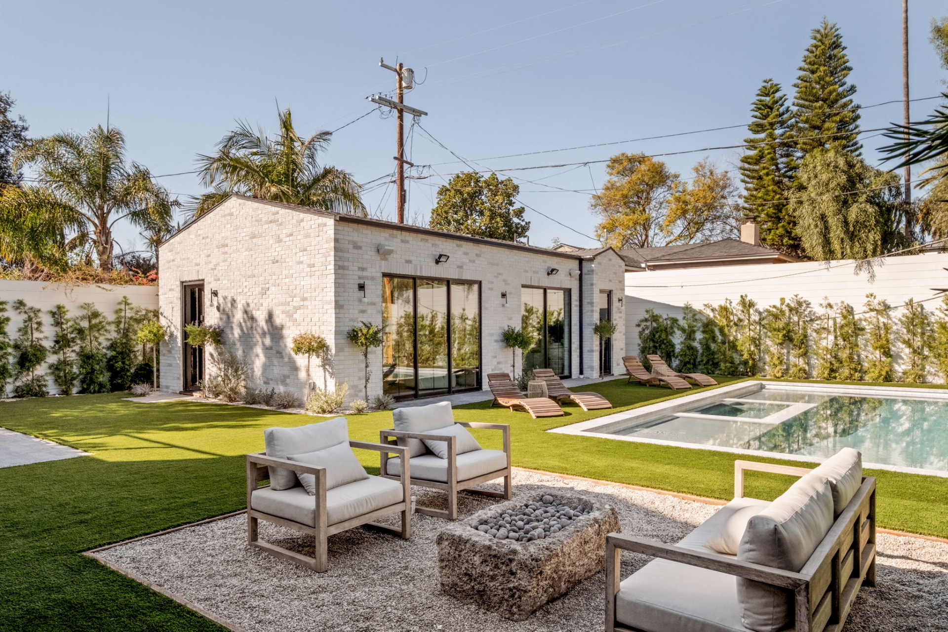 White brick ADU across the lawn with pool, spa, sun loungers and a stone fire pit seating area