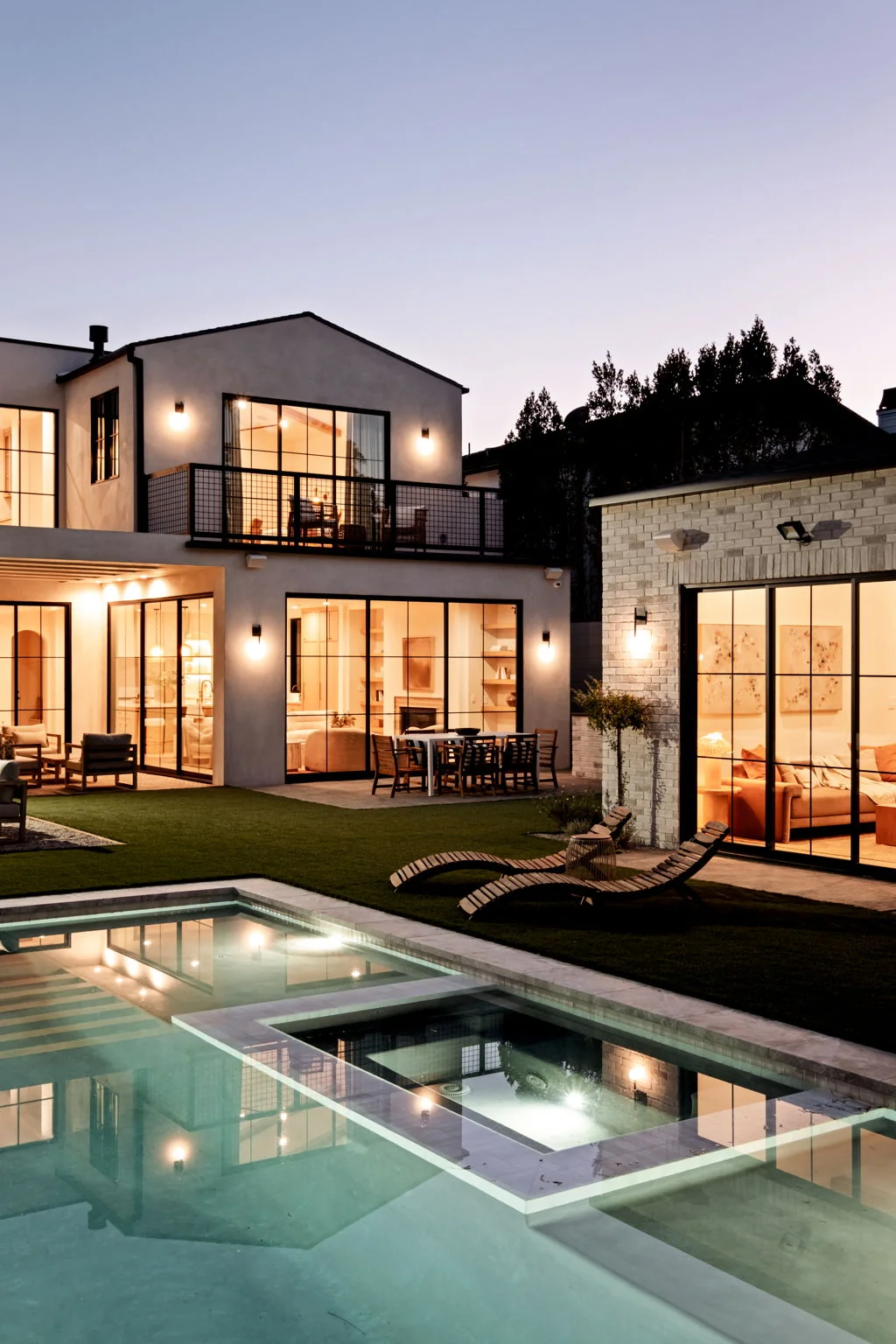 Modern Spanish residence at dusk with lit steel windows, pool and brick guest house