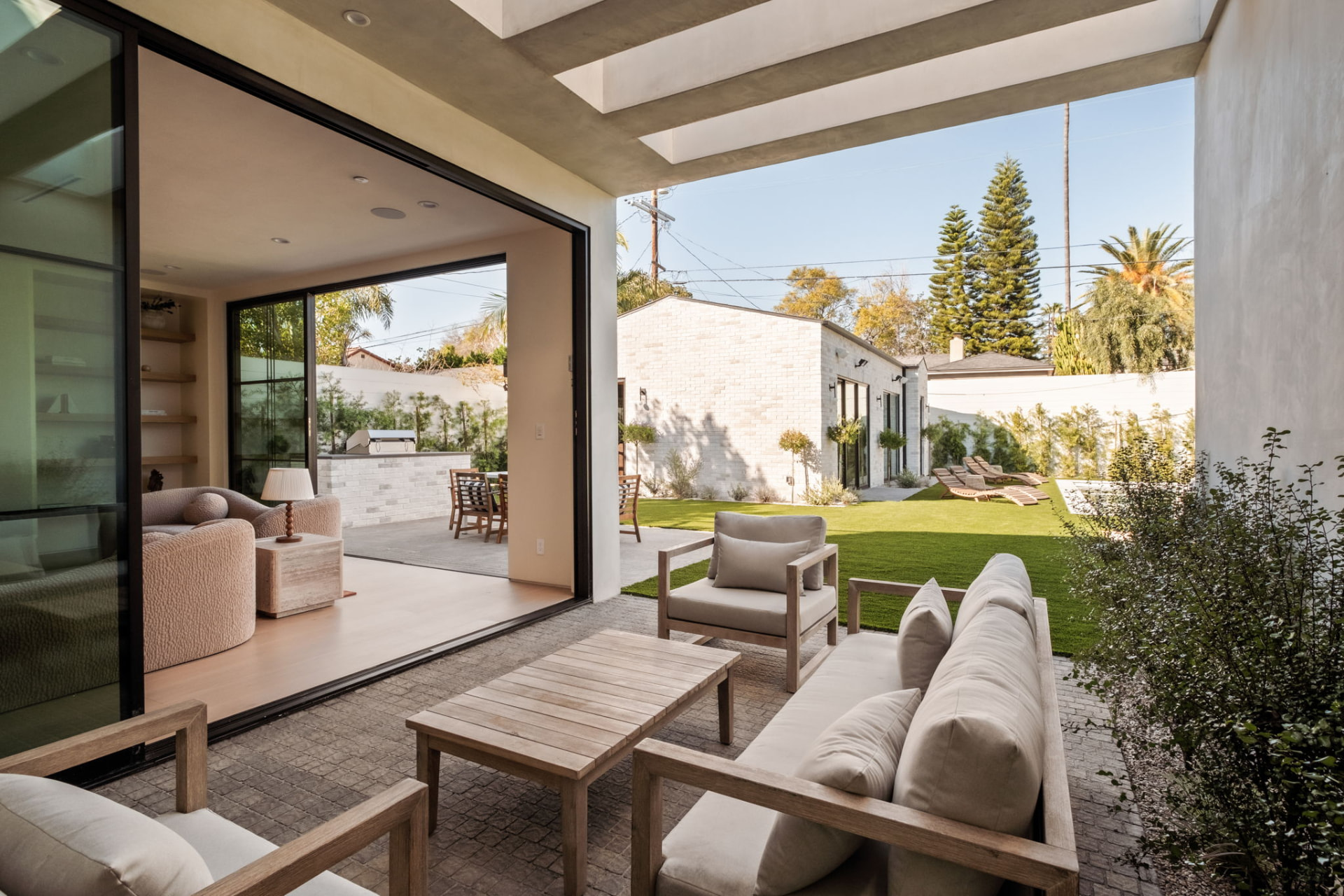 Covered loggia with a full-width pocketing glass wall opening the living room onto a teak lounge terrace, lawn and pool garden