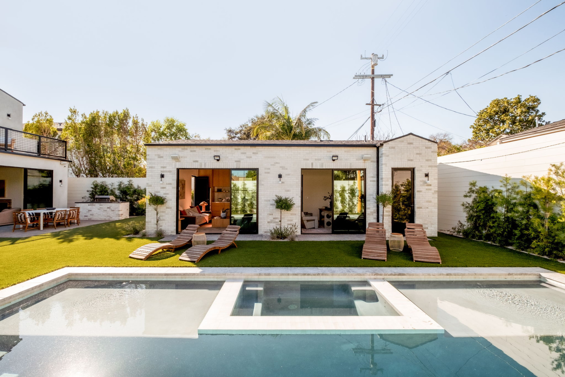 Detached white brick ADU facing the pool with sliding steel doors and lounge chairs on turf