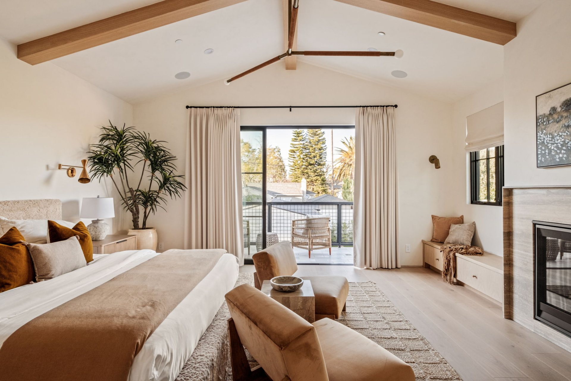 Primary suite looking out to the balcony with a stone-clad fireplace, window bench and vaulted beamed ceiling