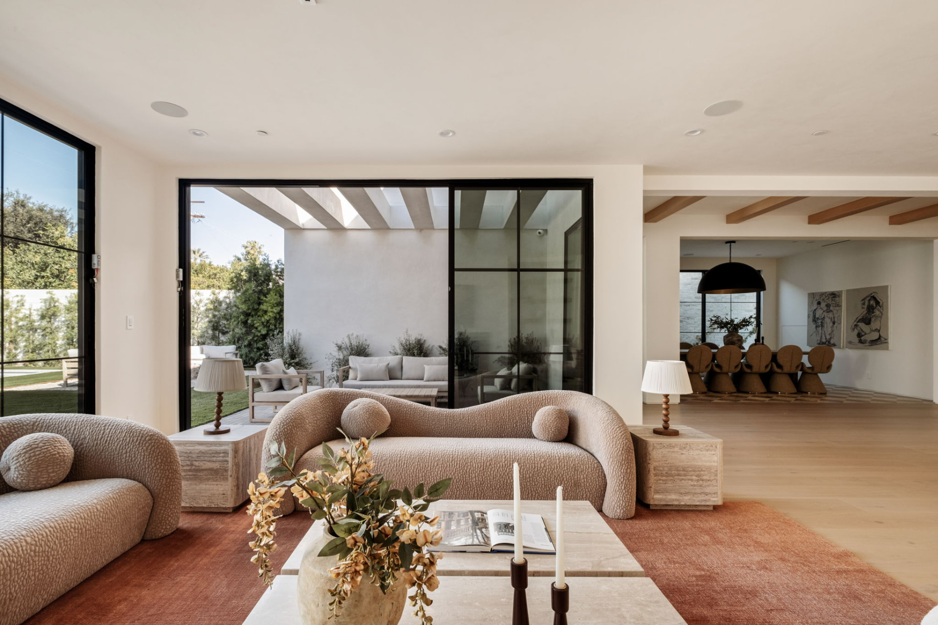 Open living room with sculptural bouclé sofas, travertine tables and full-height sliding glass doors onto a plaster courtyard