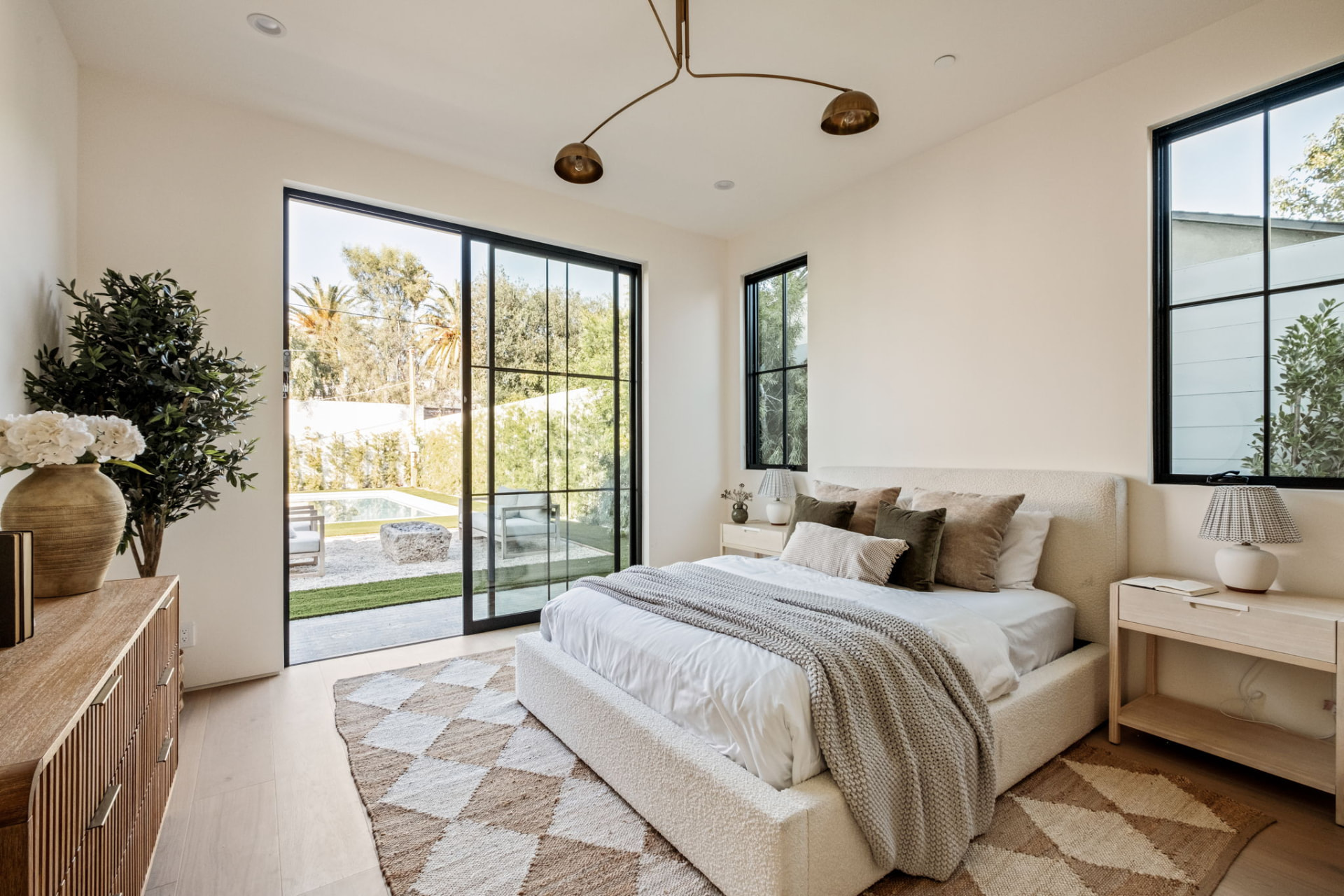 Guest bedroom with sliding steel doors onto the pool garden, a bouclé bed and a brass mobile chandelier